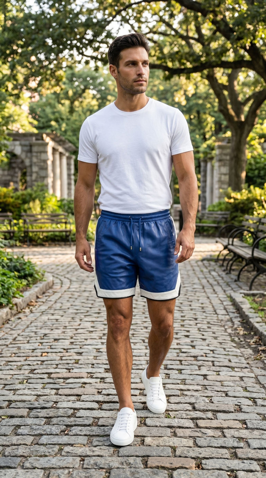 Candid shot of male model walking along cobblestone path in urban park wearing blue leather shorts and white tee.