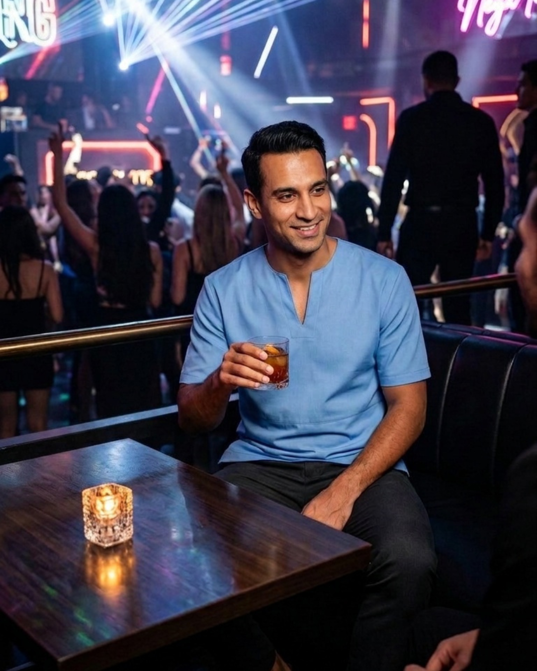 A man in a light blue V-neck leather t-shirt sits in a dimly lit nightclub , holding a drink in a glass. The background features a crowd, neon lights, and a glowing candle on a wooden table in the foreground.